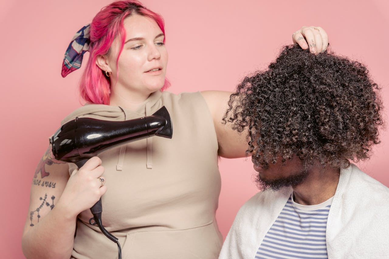 why-choose-us Female hairdresser with dyed hair drying hair of African American male model with Afro hairstyle against pink background